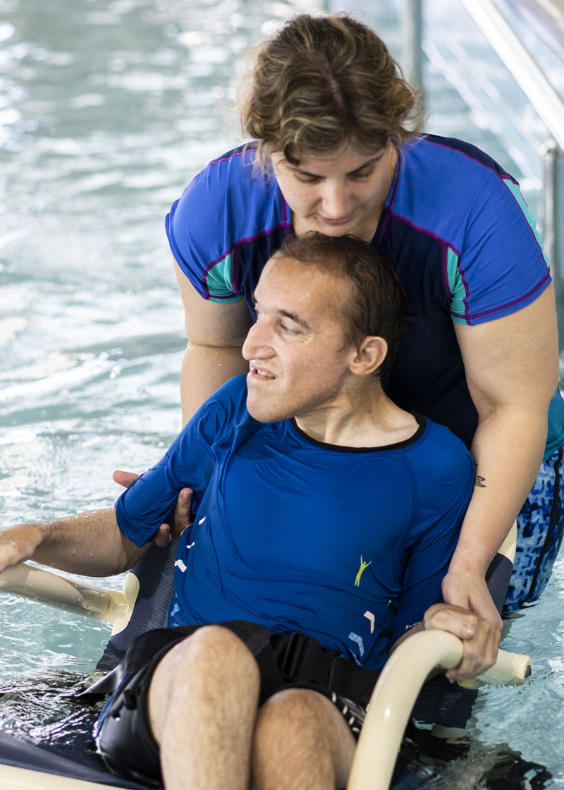 Two people in a pool, one in a wheelchair being supported by the person behind them