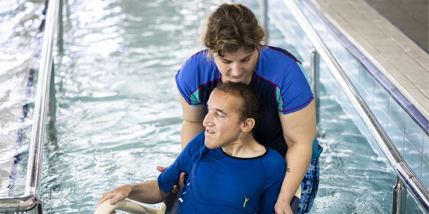 Two people in a pool, one in a wheelchair being supported by the person behind them