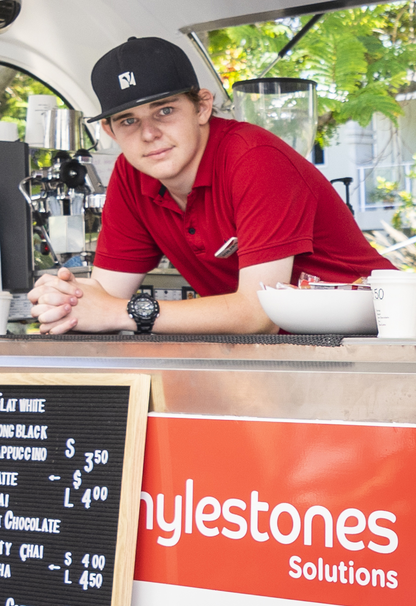 One person in a red polo shirt and a black cap leaning forwards on a bench. There is a coffee machine in the background and a Mylestones logo in the foreground