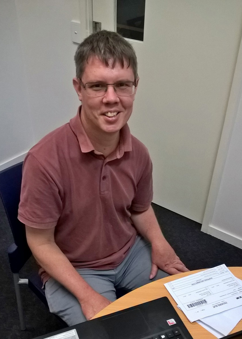 Close up image of Adam Clarke. Adam has short brown hair and is wearing black glasses and a red polo shirt. 