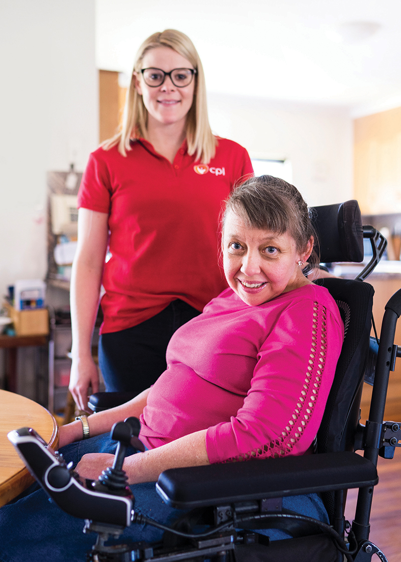 CPL Client Jenny with carer Rebecca. Jenny is in a wheelchair wearing a bright pink shirt and has short brown hair. Rebecca has shoulder-length blonde hair, black glasses and is wearing a red CPL shirt. 