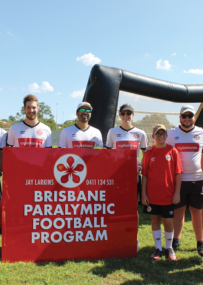 A selection of people holding a Brisbane Paralympic football program sign. The sign is red with white writing. The people are wearing white shirts with red text or red shirts with white text. the background is soccer field. 