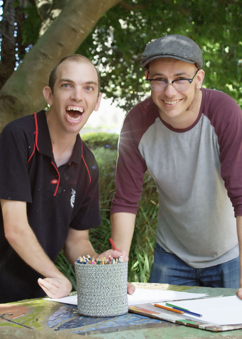 Jeremy and Chris at the park. Jeremy is wearing a grey shirt with purple sleeves, a cap and glasses. Chris is wearing black shirt and has short hair. 