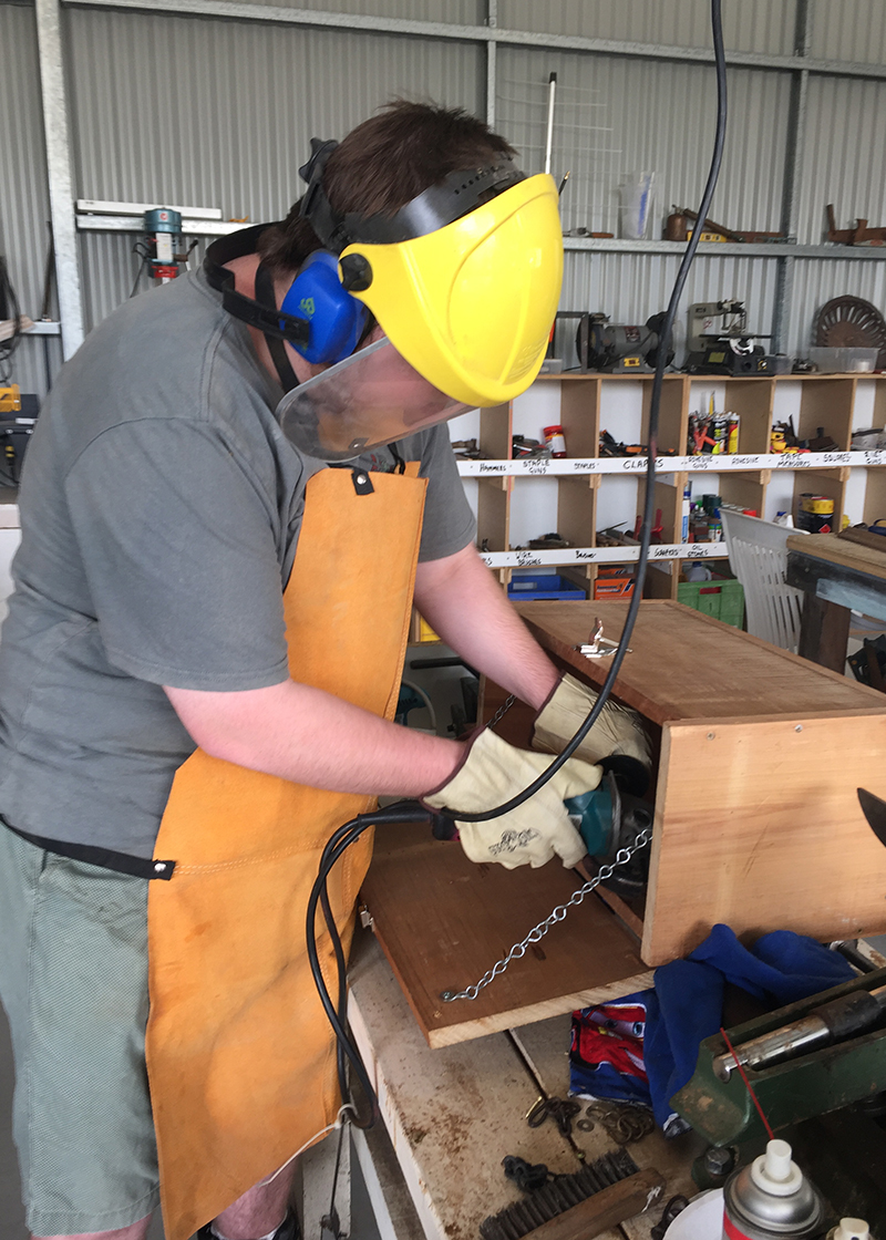 Levi working on a project at the mens shed. Levi is wearing a grey shirt and protective equipment including a face shield, ear muffs and an orange apron. 