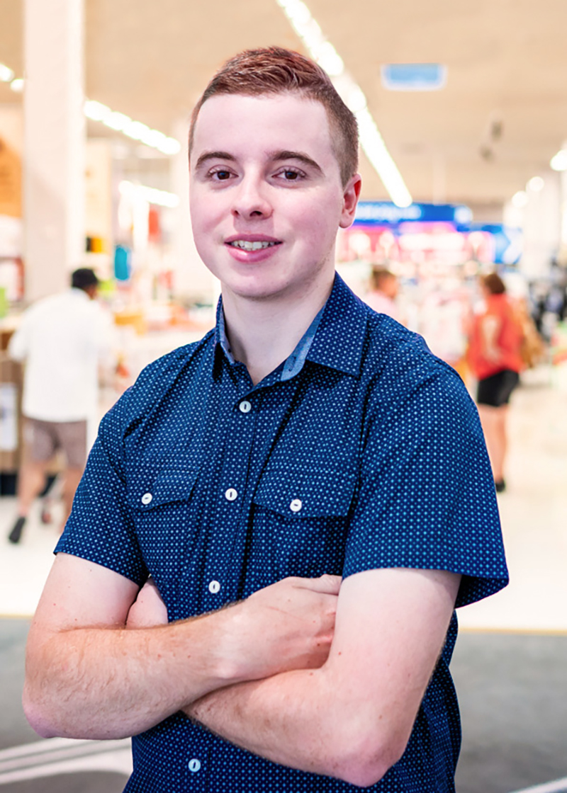Lachlan standing out front of his Kmart store. Lachlan is wearing a blue short sleeve button up shirt and has short brown hair. The background is blurred. 
