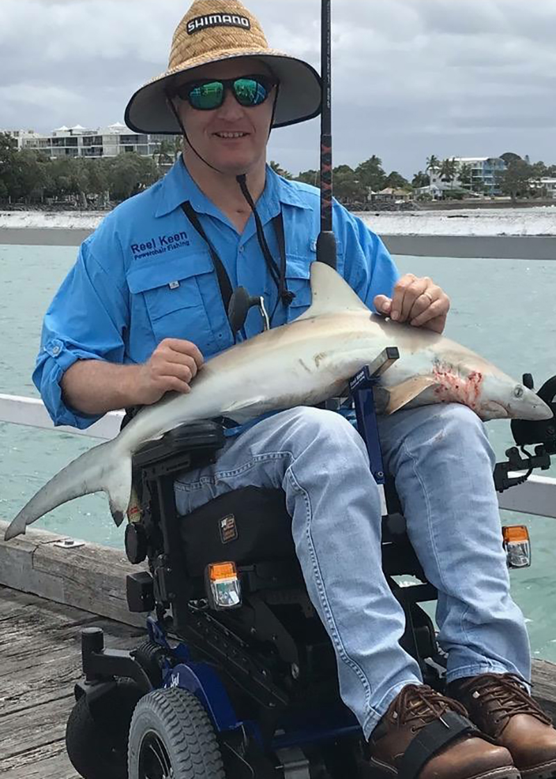 Rob Agius is holding a fish he caught on a pier. He is wearing a blue button up shirt, blue pants, black sunglasses and a straw hat. The background is of the water and sky.