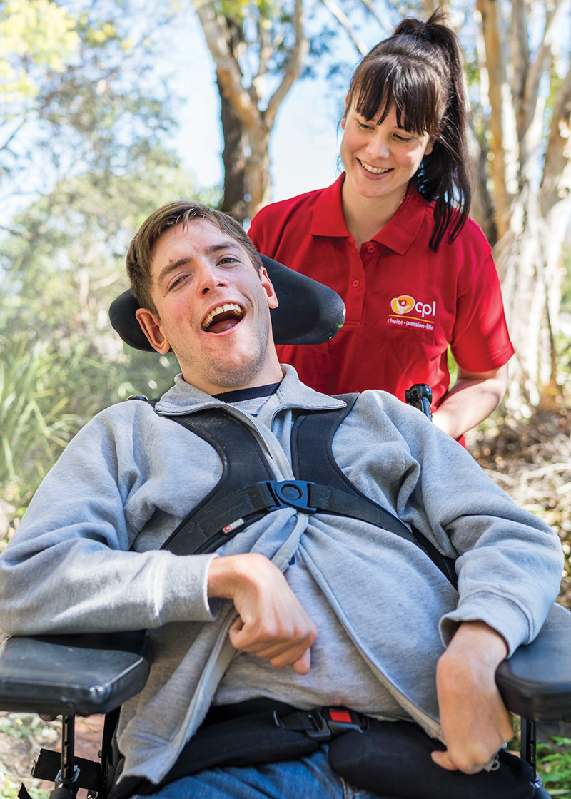 CPL client Scott Stanton with his Direct Support Worker. Scott is in a wheel chair and wearing a grey jumper. The DSW is wearing a red shirt and has long brown hair. The background is of a forest.