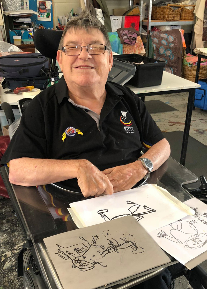Robert gallagher sitting and doing art. Robert is wearing a black polo shirt, has short grey hair and dark glasses. 