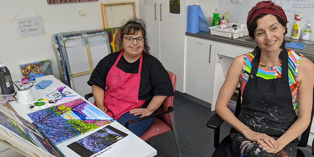 Alice Gittins sitting in the Access Arts studio with a client. Alice is wearing a green shirt with an apron with lots of paint on it. The client is wearing a black shirt with a pink apron. She is working on some painted artwork. 