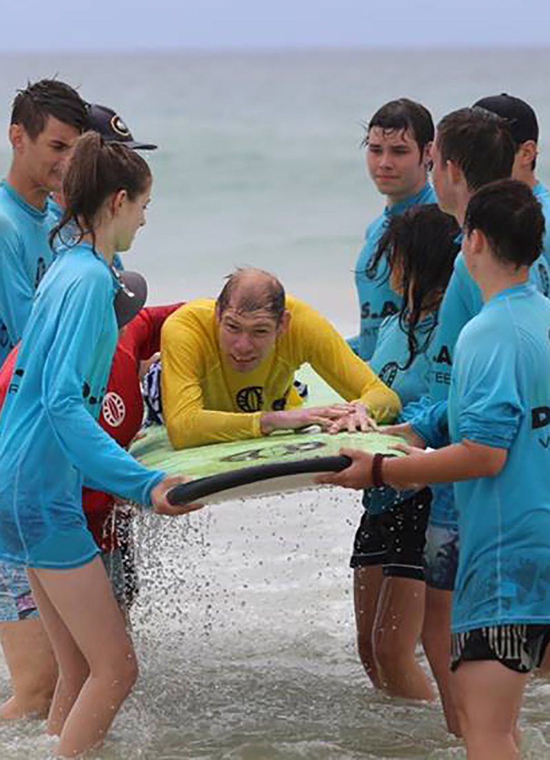 Mark learning to surf. He is wearing a yellow shirt. Mark is raised by helpers wearing blue shirts.
