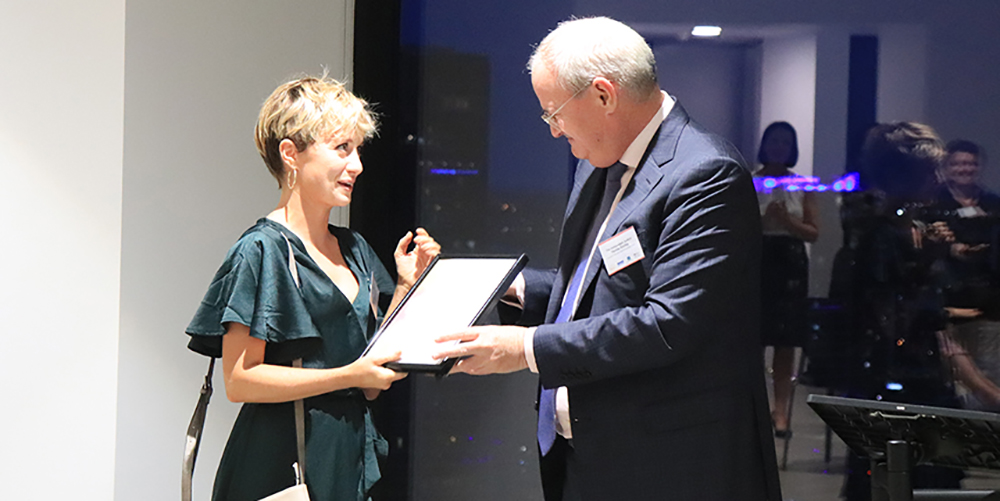 Ruby Donohoe receiving her Access Arts Achievement Highly Commended Award. Ruby has short blonde hair and is wearing a green dress. Ruby is being handed the award by a man in a suit. The man has grey hair.