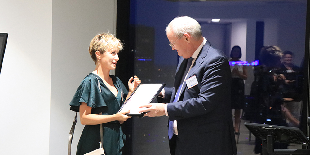 Ruby Donohoe receiving her Access Arts Achievement Highly Commended Award. Ruby has short blonde hair and is wearing a green dress. Ruby is being handed the award by a man in a suit. The man has grey hair.