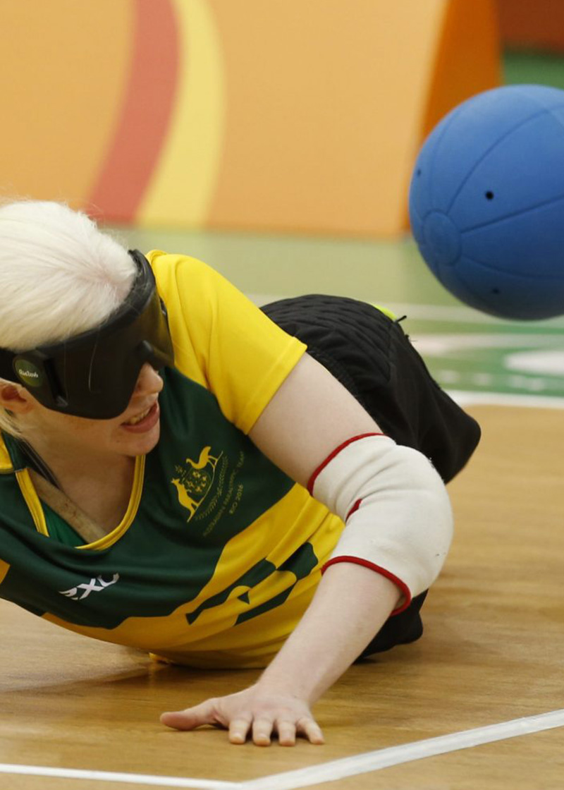 Two blindfolded Australian goalball players laying on a wooden court as a blue ball comes towards them