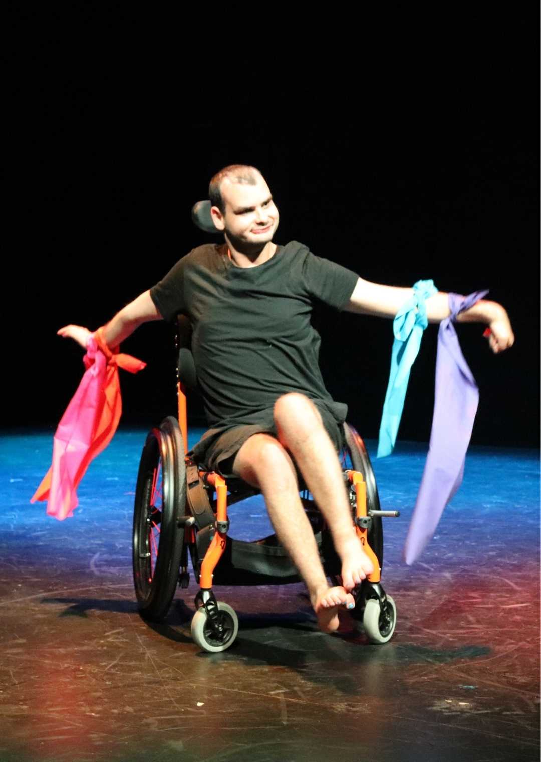 A person perfoming on stage. They have coloured ribbons tied to each of their wrists and are seated in a wheelchair