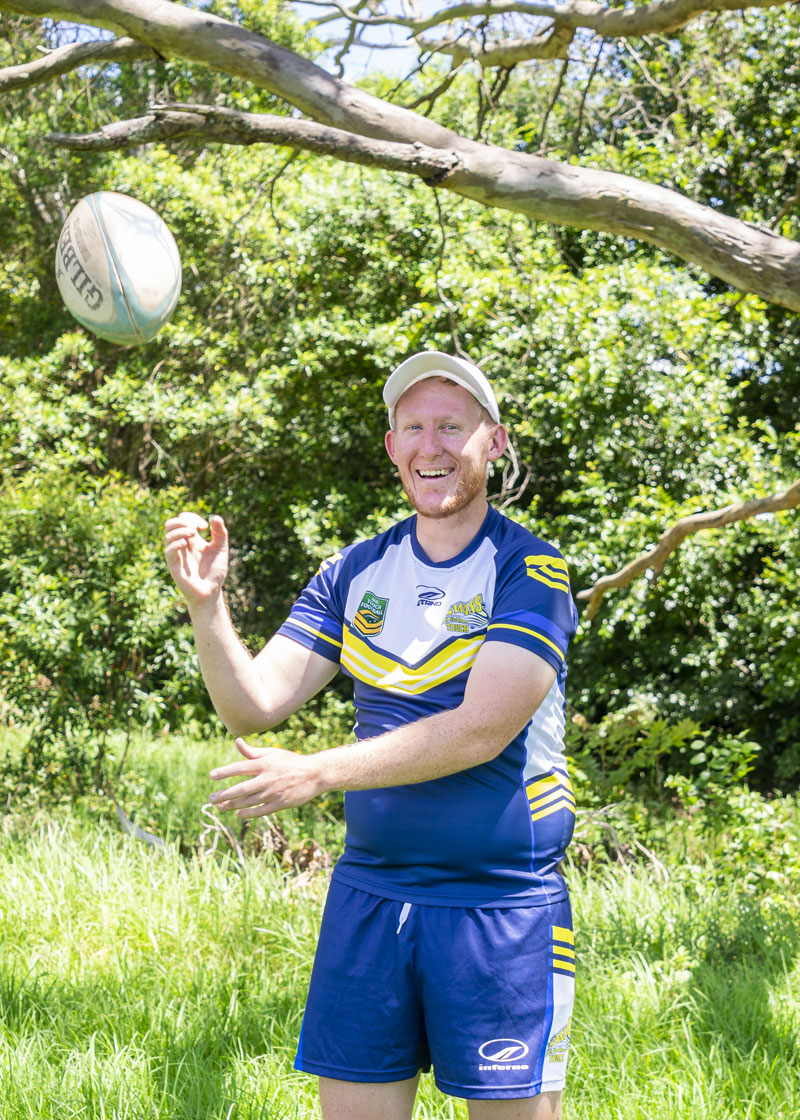 Person standing outside in a park. He is wearing a blue sports uniform and is about to catch a touch ball.