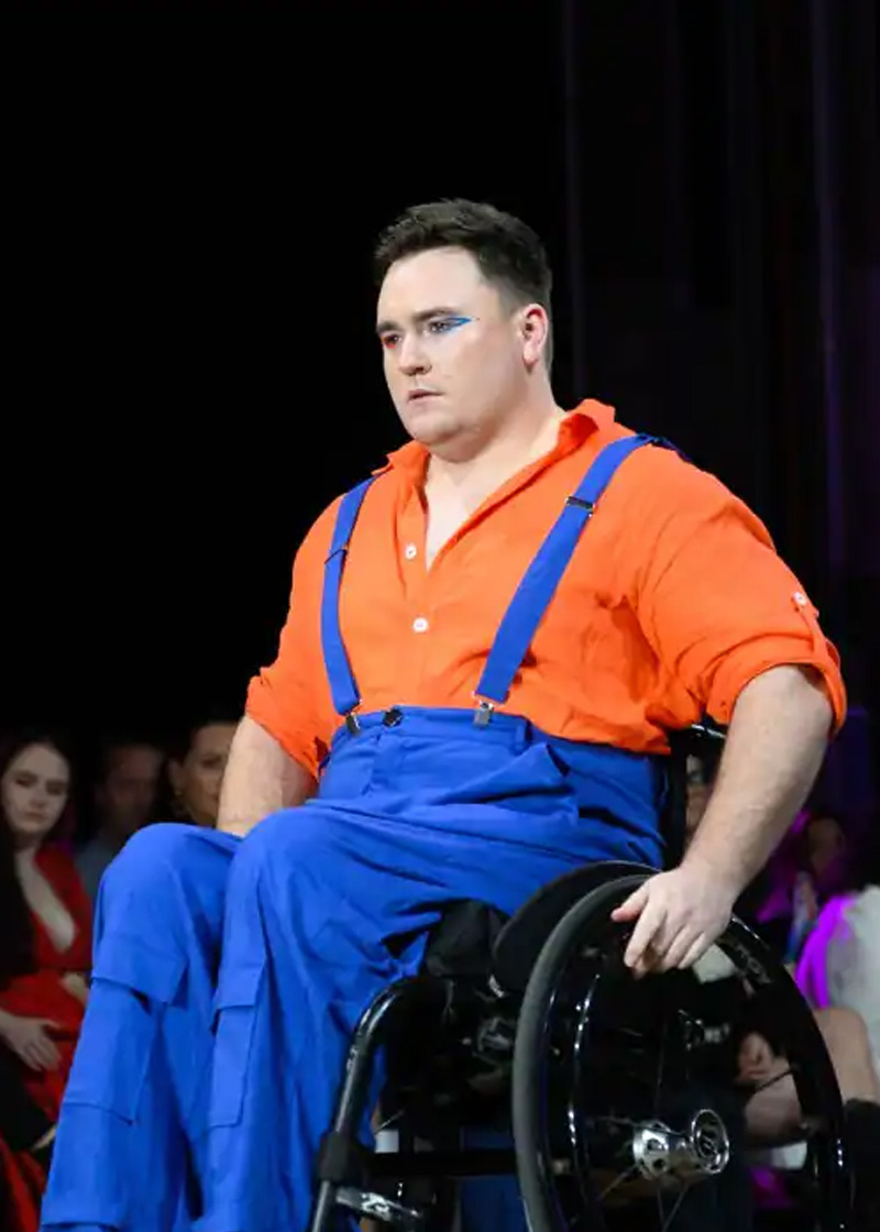 Person in a wheelchair on a fashion runway wearing a blue pants with suspenders and an orange top.