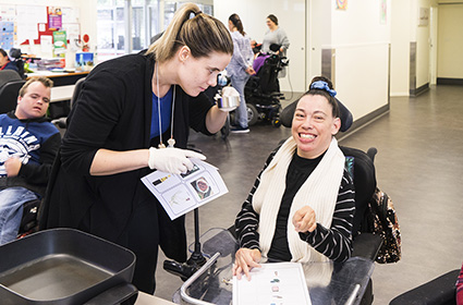 A person seated in a wheelchair and smiling as another person stands and holds a sheet with communication symbols on it towards them