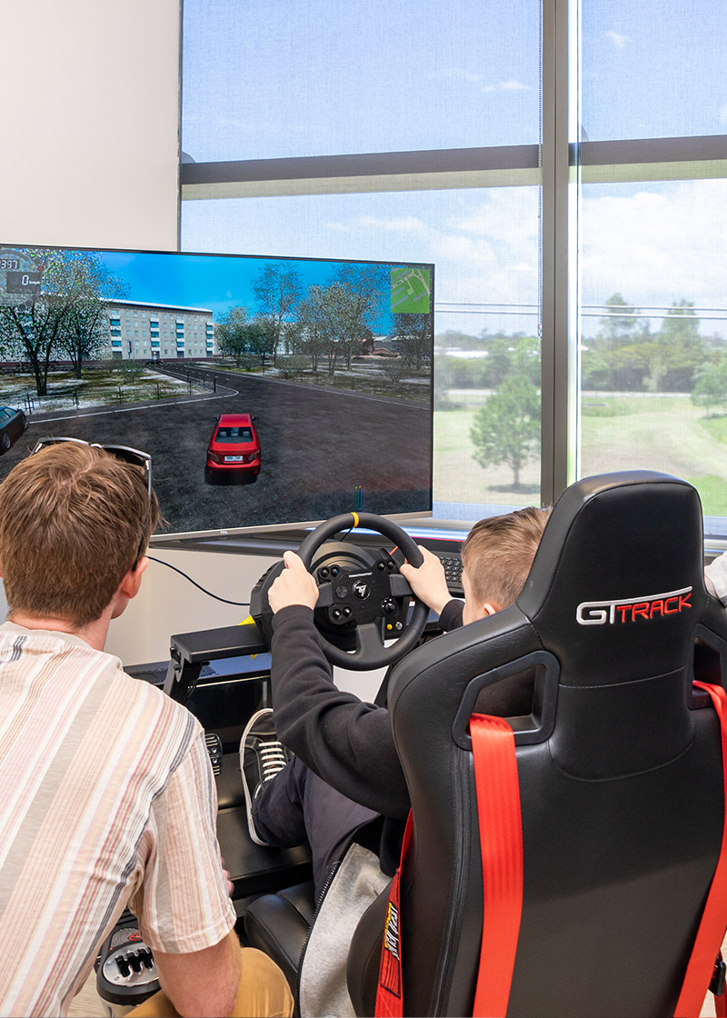 A person on the driving simulator, pictured from behind. Another person is crouched down beside them looking at the driving simulation on the screen.