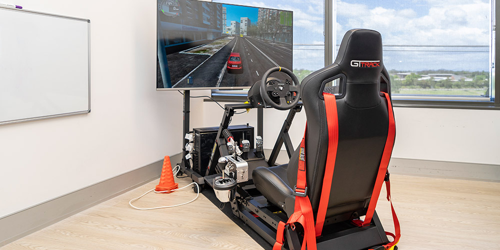 A Driver's seat in front of a steering wheel and tv screen showing a simulation of a road.