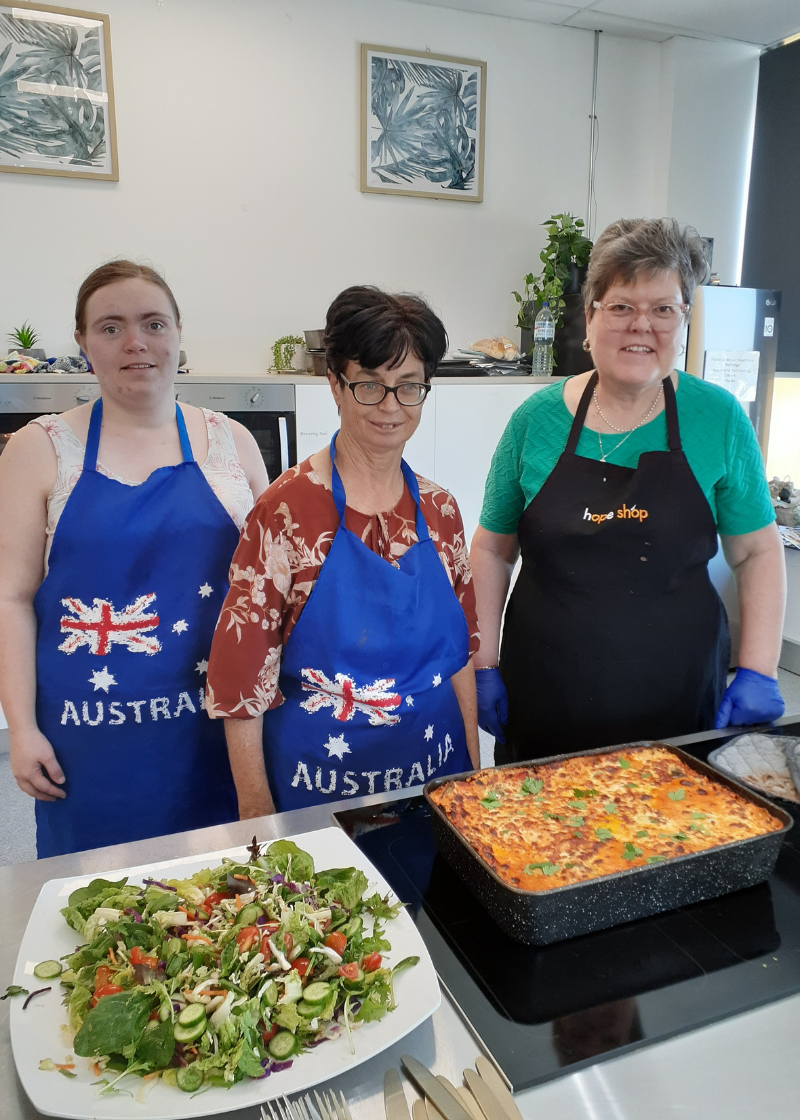 Three women standing behind a garden salad and cooked lasagne sitting on the kitchen benchtop