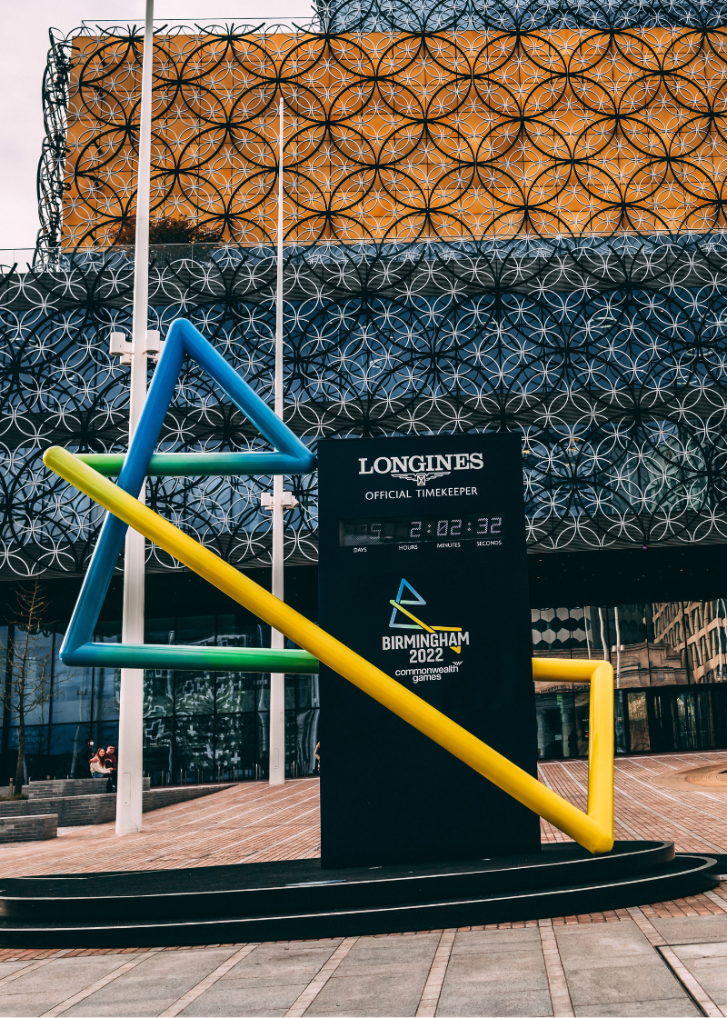 The image depicts a statue of the Birmingham 2022 Commonwealth Games. It is a variety of colourful poles coloured in a blue to yellow gradient, taking the shape of a capital B. The statue includes a black stand with a digital clock embedded with the text above saying "Longines Official Timekeeper". The statue is in front of a building with a geometric design.