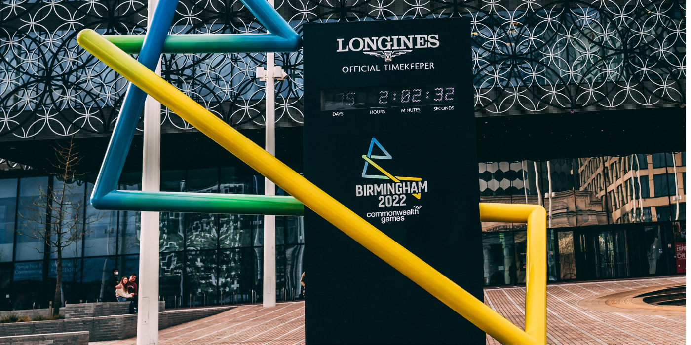The image depicts a statue of the Birmingham 2022 Commonwealth Games. It is a variety of colourful poles coloured in a blue to yellow gradient, taking the shape of a capital B. The statue includes a black stand with a digital clock embedded with the text above saying "Longines Official Timekeeper". The statue is in front of a building with a geometric design.