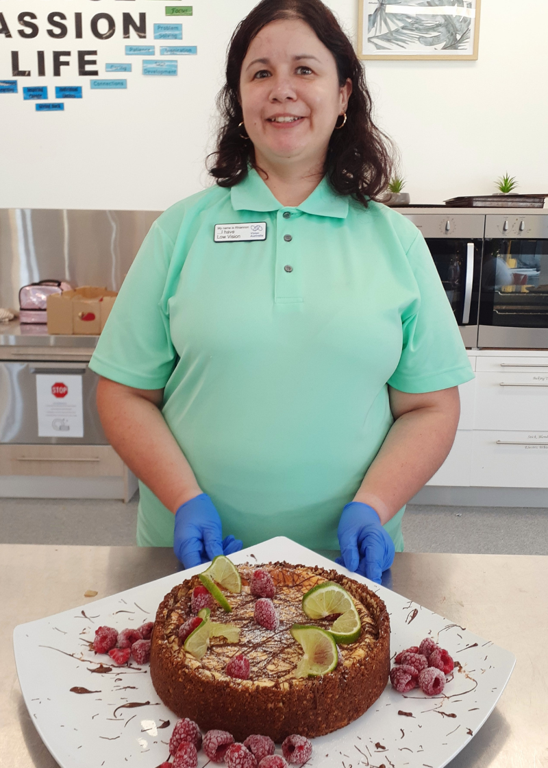 A photo of  a lady from CPL Hope Island with a light green polo shirt, blue latex gloves and long brown hair standing in a kitchen. She is holding a Key Lime Pie with dark chocolate drizzle, frozen raspberries and slices of lime on top. 