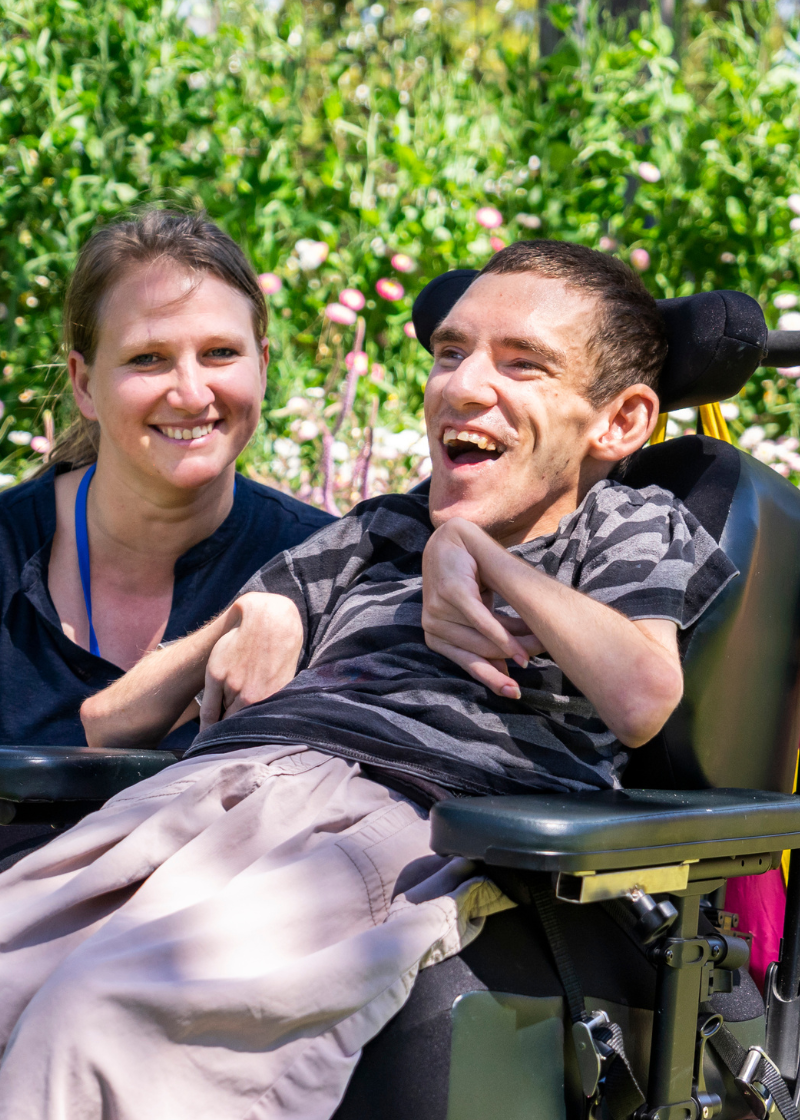 An image of a man sitting in a wheelchair with a woman kneeling beside him. They are in front of a garden of colourful plants and flowers and are smiling.