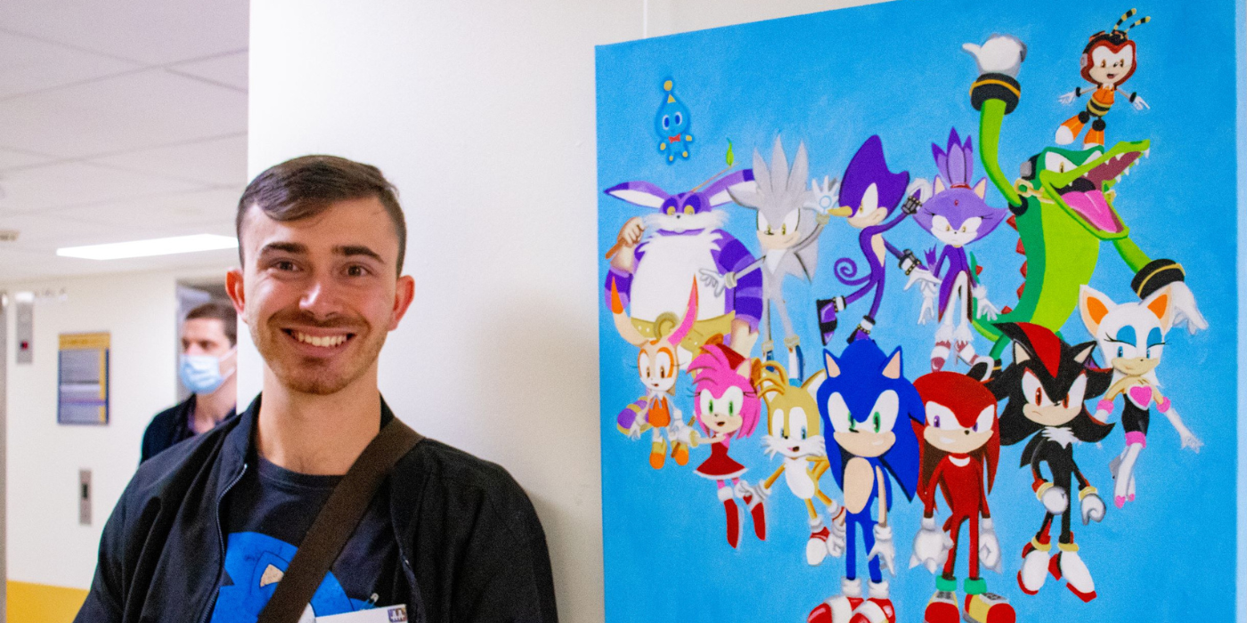 Image of a man standing next to an artwork. Man is smiling and is looking towards the camera. The artwork features different characters such as Sonic the Hedgehog. 
