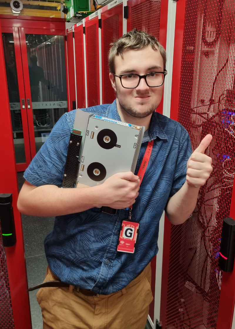 Josh is standing in front of servers holding a hard drive and smiling at the camera with his thumbs up