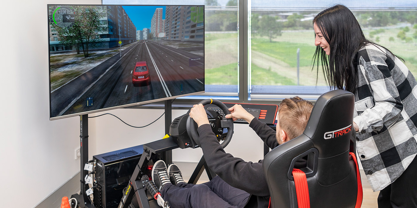 A person on the driving simulator, pictured from behind. Another person is leaning over the driver's seat smiling.