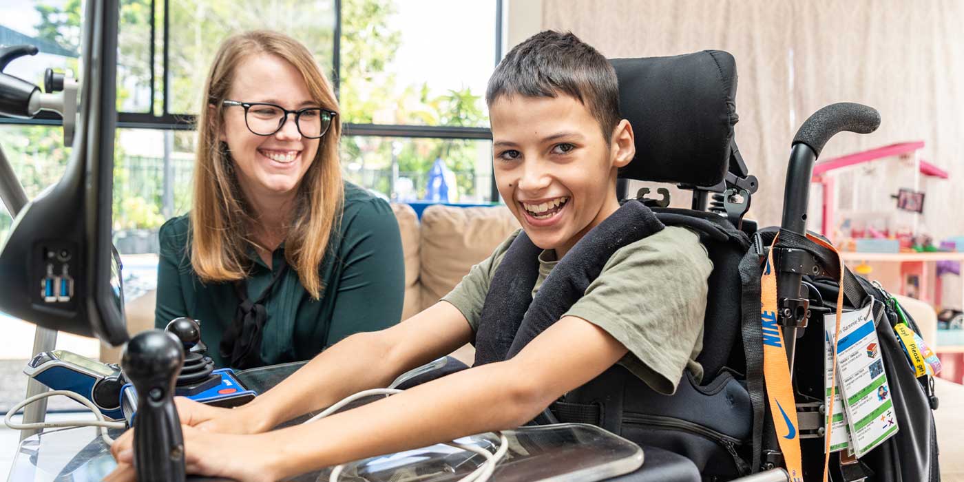 A boy sitting in a wheelchair with a lady next to him. They are both smiling.