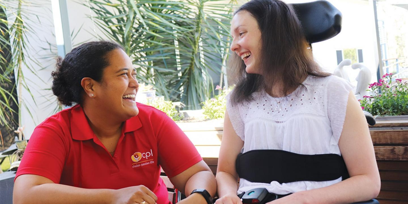 The image depicts two people sitting outside near a table, the person on the left is wearing a red, CPL branded TShirt. The person on the right is sitting in a motorised wheelchair, they are wearing a white top and their hair is brown and let down. The people are smiling at each other.